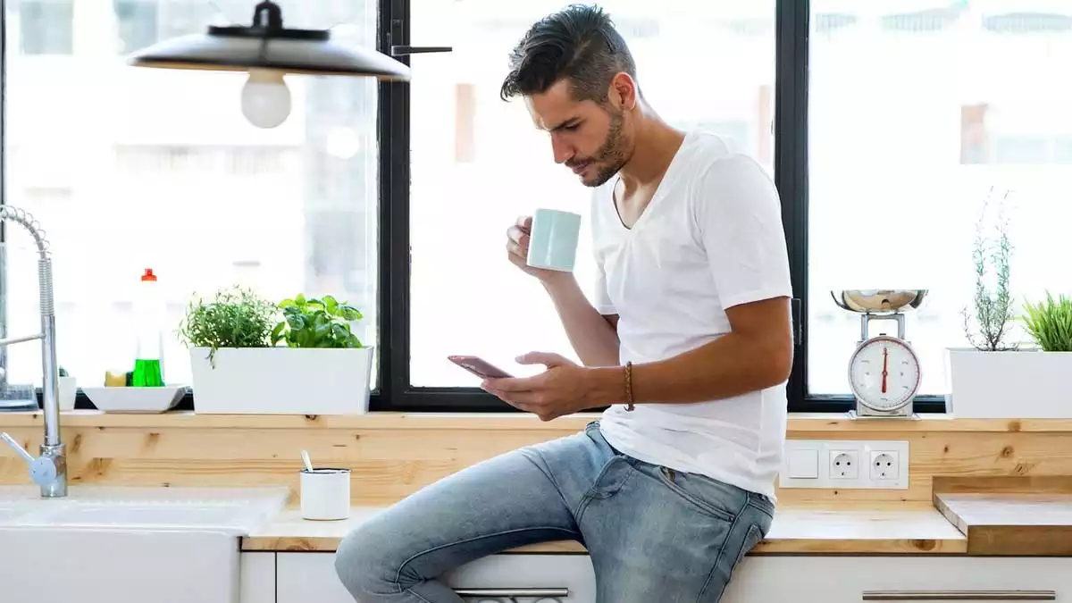 Man in a kitchen with a cup and a phone.